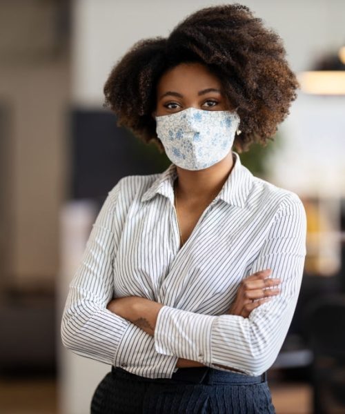 Portrait of african american businesswoman back to work at office after pandemic lockdown. Female entrepreneur with protective face mask standing alone in office with her arms crossed.